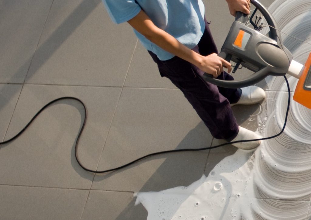 Cleaner guiding an orange floor scrubber over foamy tiles