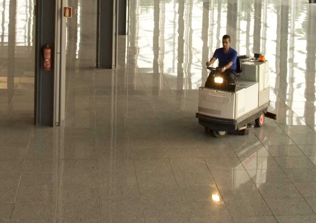 Operator driving a ride-on scrubber across a lobby floor