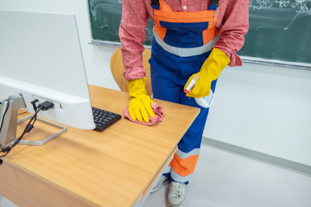 Cleaner in orange overalls wiping a classroom desk beside a computer