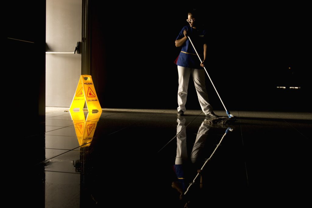 Cleaner mopping a dark reflective floor beside a caution sign