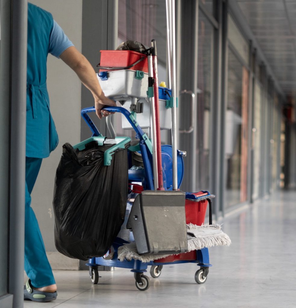Cleaner pushing a janitorial cart with a waste bag down a clinic corridor
