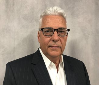 Studio headshot of Paul Figueiredo, white hair and glasses, in a dark suit jacket over an open-collar shirt